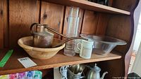 Wide view of copper watering can, wooden bowl, glass bowl, two vases, and two mugs on a wooden shelf.