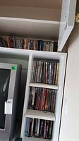 Tall white shelving unit filled with DVDs arranged in rows with some DVDs stacked vertically and horizontally. Various movie and TV show titles visible on spines.