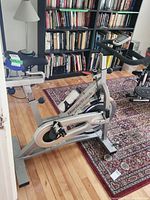 Side view of the silver NordicTrack exercise bike showing the frame, pedals, seat and handlebar setup, situated on a hardwood floor next to a red and purple patterned rug and bookshelves.