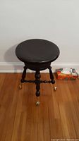 Front view of black wooden piano stool with round seat and four legs ending in claw feet on glass balls