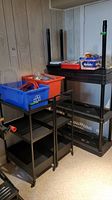 Two black metal shelving carts with blue and orange plastic bins, coiled orange extension cord, and red toolbox on black plastic shelving unit in basement corner