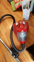 Full view of red Dirt Devil Featherlite cyclonic vacuum cleaner with hose and metal extension wand on wooden floor.