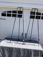 Photo of two black metal art stands with tags, positioned on a table in front of garage doors. Each stand has a triangular frame and horizontal bar for holding artwork.