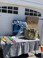 Camping cot, cardboard box, Yeti cooler, and kites displayed on table covered with gray cloth outdoors in front of garage door.