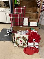 Photo showing assorted Christmas decor including plaid throw pillows, decorative holiday pillows, and several red velvet Santa sacks. Some socks also visible on floor mat.