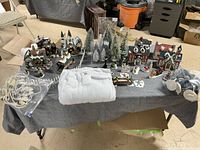 Wide view of the Christmas village display on a table showing multiple buildings, figurines, pine trees, and fake snow accessories organized over a grey tablecloth.