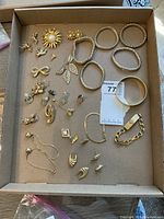 Top view of assorted gold-tone costume jewelry in a cardboard tray including bracelets, earrings and pins.