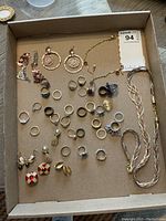 Photo showing assorted rings, earrings, chain necklace, braided cord necklace, and a bracelet arranged in a box.