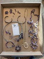 Full view of assorted costume jewelry including necklaces with purple stones, gold-tone pendant necklace, silver choker, bracelets, various purple earrings, and a large pin.