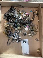 Top-down photo of a cardboard box containing a tangled assortment of costume jewelry including chains, bracelets, earrings, and pins.