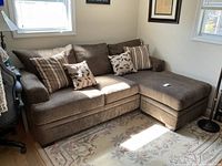 Brown fabric L-shaped sectional couch with multiple decorative pillows, placed in a corner with windows providing natural light.