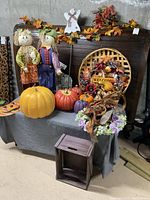 Two fabric scarecrow dolls, multiple artificial pumpkins, circular woven basket wreath with fall decorations, floral arrangement, and small wooden crate on and around a gray cloth-covered table.
