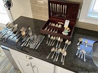 Overhead photo of assorted silver plate flatware including forks, knives, spoons, serving spoons, ladles, and measuring cup spread out on dark countertop with flatware box with knives in background