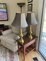 Two matching brass-colored table lamps with gray fabric bell-shaped shades on a wooden side table next to an upholstered armchair and TV.