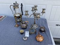 Mixed metal decorative items including silver plate candelabra, glass carafe, copper teapot, salt dishes, magnifying glass, and brass candlesticks arranged on a grey background.