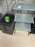 Overview of 3-tier glass shelving unit, black 3-drawer filing cabinet, wire letter holder, and pencil cup grouped together on floor.