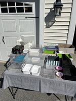 Various plastic storage containers and bins in clear, white, black, and colorful varieties arranged on a table outdoors in sunlight.