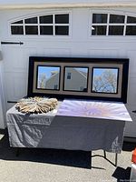 Full view of all three items arranged on table outside against garage door: large triple-paneled mirror, driftwood sunburst, floral photo print