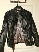 Front view of black Harley Davidson women's leather jacket hanging on hanger, showing front zipper partially open and orange piping detail on right front side. The leather has a slightly worn look reflecting light, and the interior lining is visible.