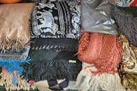 Top down view of a pile of various scarves and shawls showing different colors, patterns, and textures including beige, black, orange, and grey scarves with fringes and some patterned items.