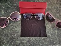 Three pairs of ladies sunglasses arranged on a dark marbled surface with a red Guess branded case and black cleaning cloth.