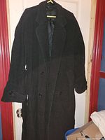 Full front view of men's black long wool coat with buttons and lapel collar hanging on hanger against door, showing overall condition and style.