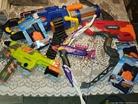 Photo shows entire lot of Nerf toy guns and accessories arranged on a table with a lace cloth surface.