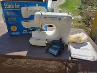 Full view of white Stitch Art sewing machine with model and brand showing on box behind. Accessory box and cords visible.