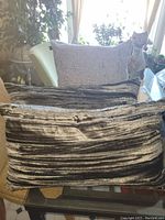 Three throw pillows displayed on a glass table and chair, one grey square pillow in the back and two silver textured rectangular pillows in front.