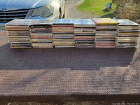 Large stacks of various music CDs in jewel cases shown outdoors on a cloth surface.