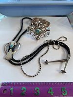 Photo showing bolo tie with silver turquoise centerpiece along with two silver cuff bracelets with turquoise stones, placed on a flat surface with ruler showing size