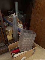 Box containing a collection of various colored taper candles and a glass rectangular dimpled candle holder, along with a tall clear glass vase and other glass items in the background.