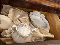 Photo showing lidded sugar bowl, plates, and bowls with floral trim, and folded beige table linens.