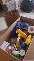 Photo of box containing multiple cleaning supplies such as spray bottles, Swiffer wipes, mop, and cleaning bottles.