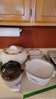 Overview of kitchen countertop showing all items in lot including covered white round insulated bowl, brown ceramic pot, two CorningWare dishes (square and rectangular)
