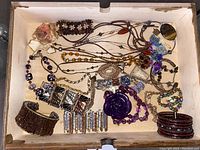 Photo showing assortment of gold-toned jewelry including bracelets, cuffs, pendants, pins, and necklaces in a wooden tray with various colors and materials.