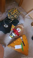 Photo showing cheese-head cutting board, black Packers tote bag, pot holders, Bobblehead figure, and glimpse of sweatshirts and t-shirts in pile.