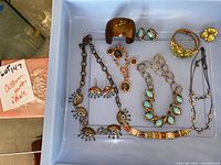 Overhead view of assorted costume jewelry including eye-themed necklace and earrings, cuff bracelet, coin charm bracelet, green leaf-themed jewelry, beaded necklace, floral earrings, and a stone container with box