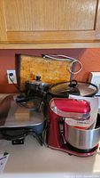 Photo showing red Dash Everyday Stand Mixer and Rival slow cooker with glass lid placed on counter with warming tray and electric fry pans.