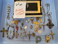 Top-down photo of all religious jewelry laid out in a blue plastic bin including the jewelry box, rosary, crucifixes, and various charms and pendants
