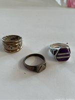 Photo of three rings including a wide band with star and sunburst motifs, a signet ring, and a purple and gold tone decorative ring.