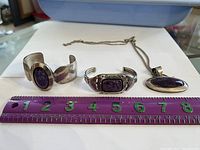 Three items displayed together: two sterling silver cuff bracelets and one pendant with a chain. Purple stones are centrally set in each piece. The items are placed on a white background with a purple ruler for scale showing approximately 7 inches in length.