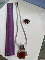 Silver necklace with flat chain and large oval red gemstone pendant next to a purple ruler for size reference showing necklace approximately 15 inches long