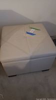Front of white square ottoman with stitched panel design on top, showing top surface and overall shape.