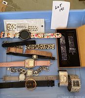 Photo of nine watches and watch bands arranged on a blue tray including Samsung smartwatch with floral band and various analog watches and bands.