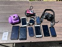 Overview photo showing all devices from above on a wooden surface, including multiple smartphones, cameras, an instant film camera, and charger cables.