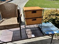 Vintage wicker chair with woven back and seat on brown metal frame alongside two-drawer wicker storage table on metal frame with seat cushion on the ground.