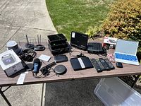 Photo showing various electronics arranged on table including a boxed wireless speaker, headphones, microphones, remotes, routers, a laptop, and multiple cables.