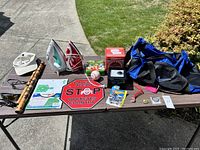 Photo shows assorted household items laid out on a brown table outside on pavement. Items include two irons, a duffel bag, wooden flute, sunglasses, various small objects like a baseball, wire cutter, decorative box, and novelty sign.