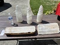 Five large selenite crystals displayed on a table outdoors. Three are tower-shaped polished crystals and two are flat raw slab crystals. A water bottle is shown for scale.